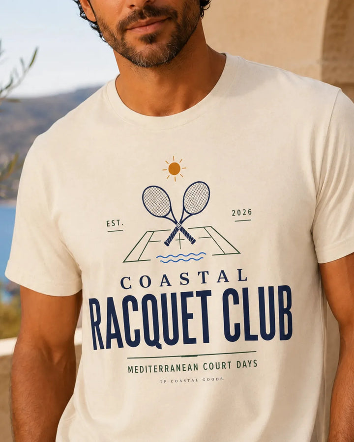 Man wearing a Coastal Racquet Club T-shirt with a Mediterranean court days graphic print in a sunlit coastal setting.