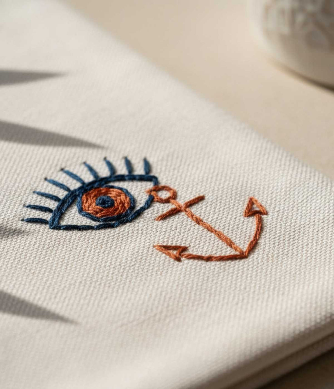 Close-up of embroidered eye and anchor in blue and terracotta thread on beige canvas fabric.