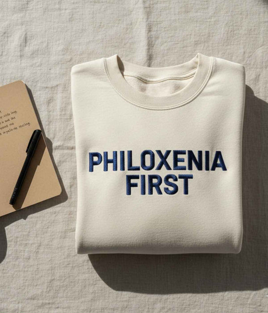 Cream sweatshirt with bold navy embroidery reading “Philoxenia First,” placed beside a notebook in warm natural light.