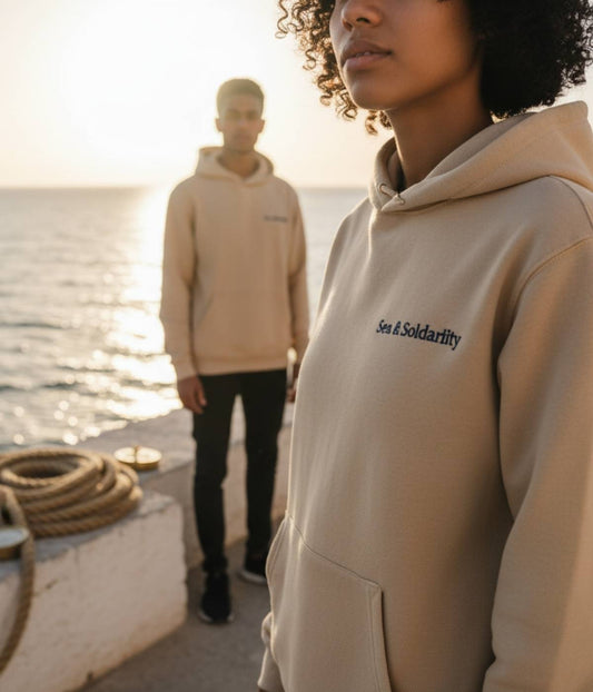Two people wearing beige “Sea & Solidarity” hoodies standing on a seaside pier at sunset.