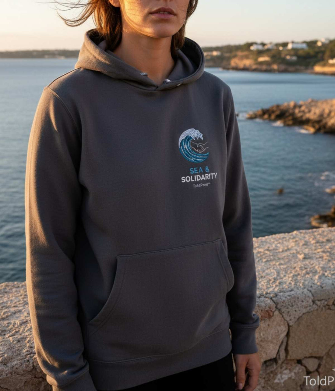 Person wearing dark gray hoodie with blue wave embroidery and “Sea & Solidarity” text overlooking the sea.