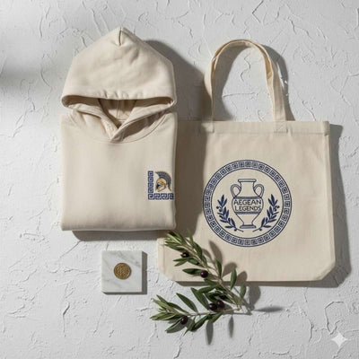 Flat-lay of Greek-inspired embroidered hoodie and tote bag with subtle ancient Greek motifs. White plaster background, olive branch, marble accent, and soft Aegean light.