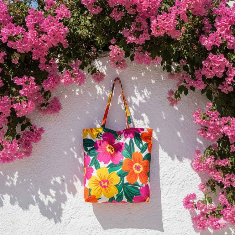 Tote bag with bright floral print on white plaster wall background. Mediterranean light, pink bougainvillea around, summery vibe.
