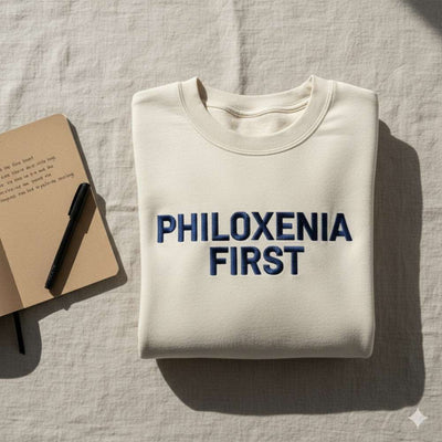 Flat-lay of activist-style sweatshirt with embroidered slogan “Philoxenia First.” Natural light, notebook and pen nearby, minimalist activist aesthetic.