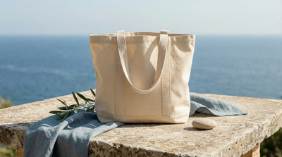 a natural cotton canvas tote bag placed on a sunlit stone surface near the sea, with subtle coastal elements
