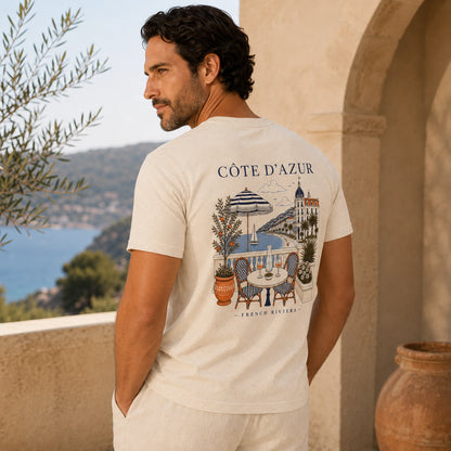 A man wearing a cream-colored t-shirt with a detailed graphic print of a French Riviera 'Côte d'Azur' scene on the back, standing on a sunny stone balcony overlooking the sea.