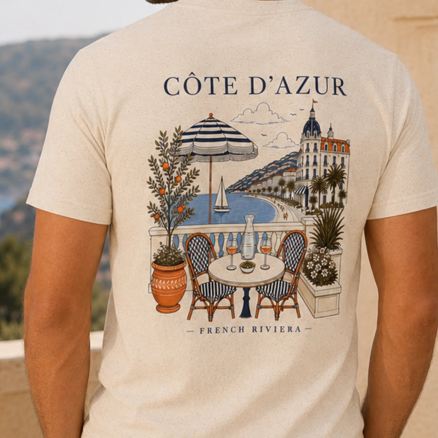 TP HERO: A close-up of a person wearing a light beige t-shirt, showing the back of the shirt with a detailed graphic print of a French Riviera seaside scene. The print features the text 'CÔTE D'AZUR' and 'FRENCH RIVIERA' with an illustration of a balcony table, striped umbrella, palm tree, and coastal architecture.