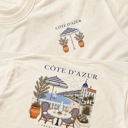 Close-up of a cream-colored t-shirt showing the front and back designs. The back features a detailed illustration of a coastal balcony scene labeled 'CÔTE D'AZUR' in navy blue text, while the front has a smaller version of the same graphic.
