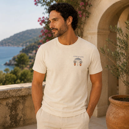A man wearing a cream-colored short-sleeve t-shirt with a small graphic print on the left chest, standing on a sunny outdoor terrace with a coastal view in the background.
