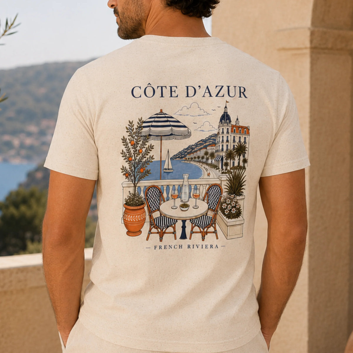 TP HERO: A close-up of a person wearing a light beige t-shirt, showing the back of the shirt with a detailed graphic print of a French Riviera seaside scene. The print features the text 'CÔTE D'AZUR' and 'FRENCH RIVIERA' with an illustration of a balcony table, striped umbrella, palm tree, and coastal architecture.