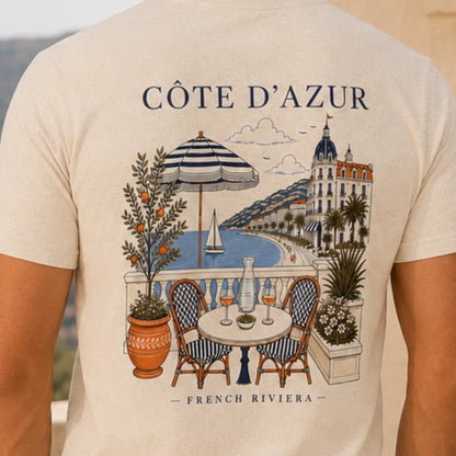 TP HERO: A close-up of a person wearing a light beige t-shirt, showing the back of the shirt with a detailed graphic print of a French Riviera seaside scene. The print features the text 'CÔTE D'AZUR' and 'FRENCH RIVIERA' with an illustration of a balcony table, striped umbrella, palm tree, and coastal architecture.