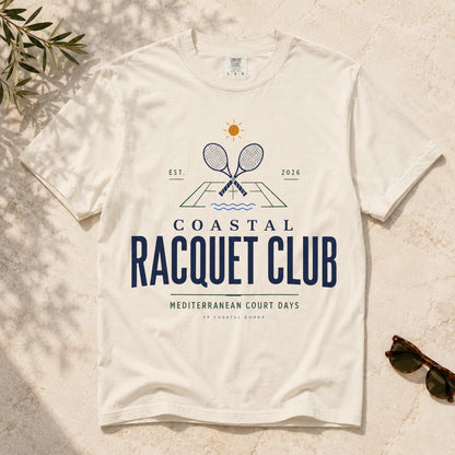 White t-shirt with 'Coastal Racquet Club' design on a textured surface with sunglasses and leaves.