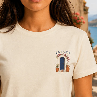 A close-up of a woman wearing a cream-colored t-shirt with a small graphic on the left chest depicting a blue door, potted plants, and the word 'ESPAÑA' under a floral archway, set against a blurred outdoor background.