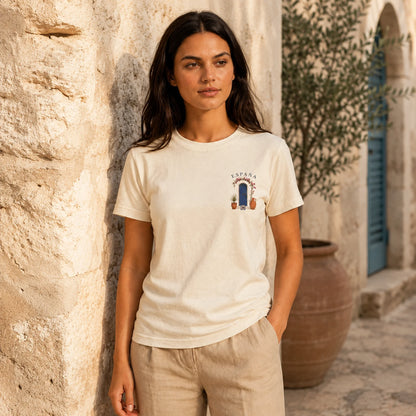 A woman stands outdoors against a sunlit stone wall wearing a light cream short-sleeve T-shirt with a small chest graphic and beige pants.