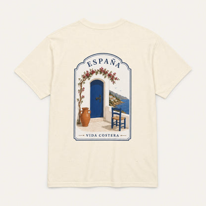An off-white short-sleeve t-shirt with a large graphic print on the back depicting a Mediterranean scene with a blue door, blue sky, and the text 'ESPAÑA' and 'VIDA COSTERA'.