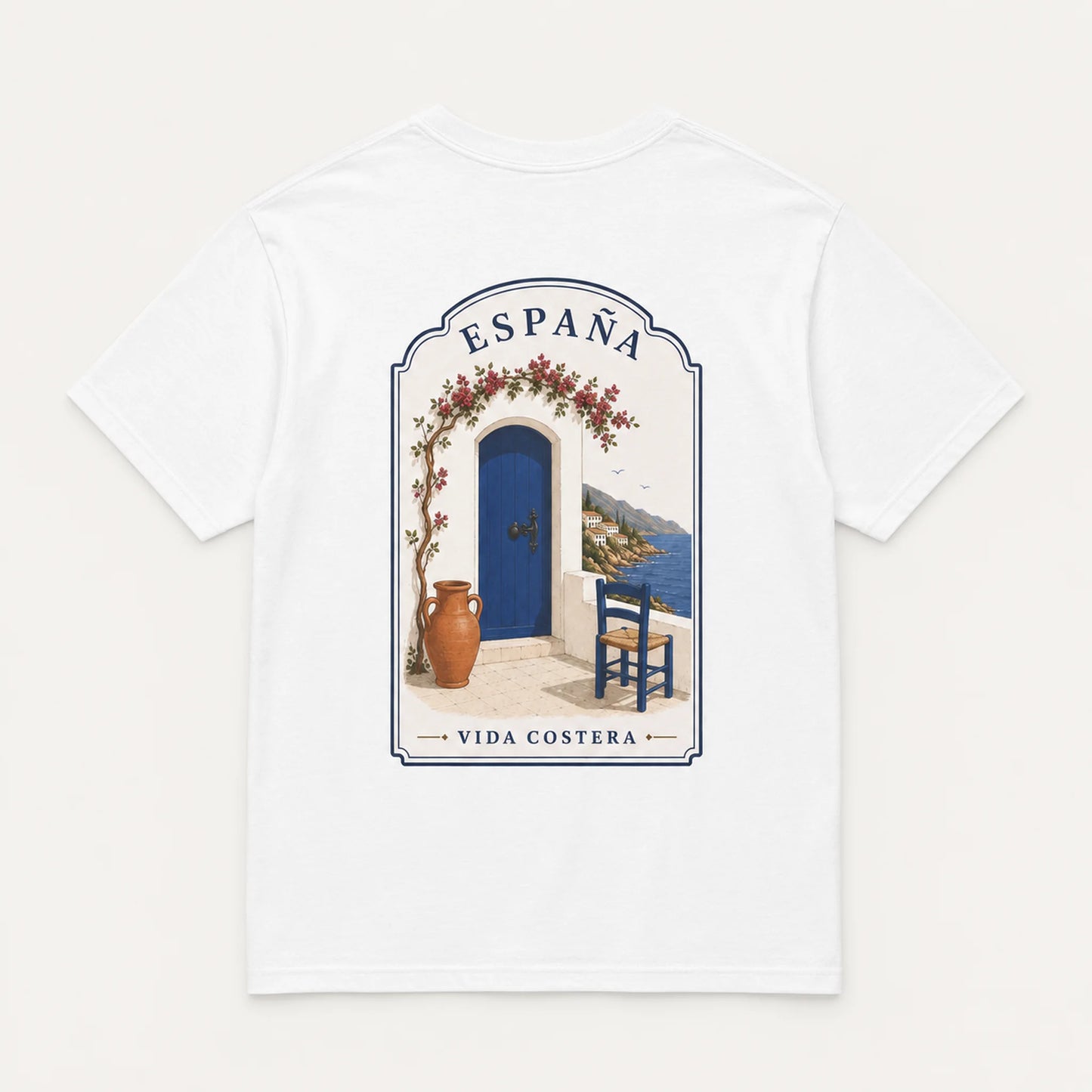A white short-sleeve crewneck t-shirt featuring a central graphic print of a Mediterranean-style doorway with a blue door, terracotta vase, blue chair, and the text 'ESPAÑA' and 'VIDA COSTERA'.