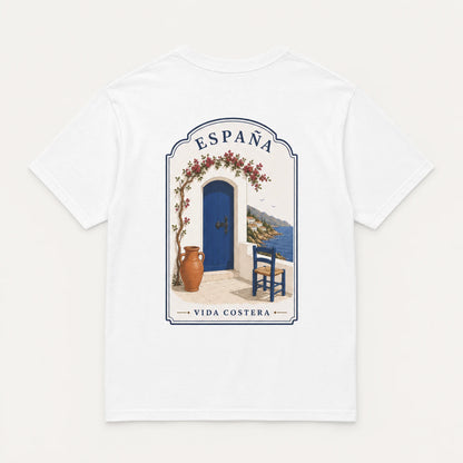 A white short-sleeve crewneck t-shirt featuring a central graphic print of a Mediterranean-style doorway with a blue door, terracotta vase, blue chair, and the text 'ESPAÑA' and 'VIDA COSTERA'.