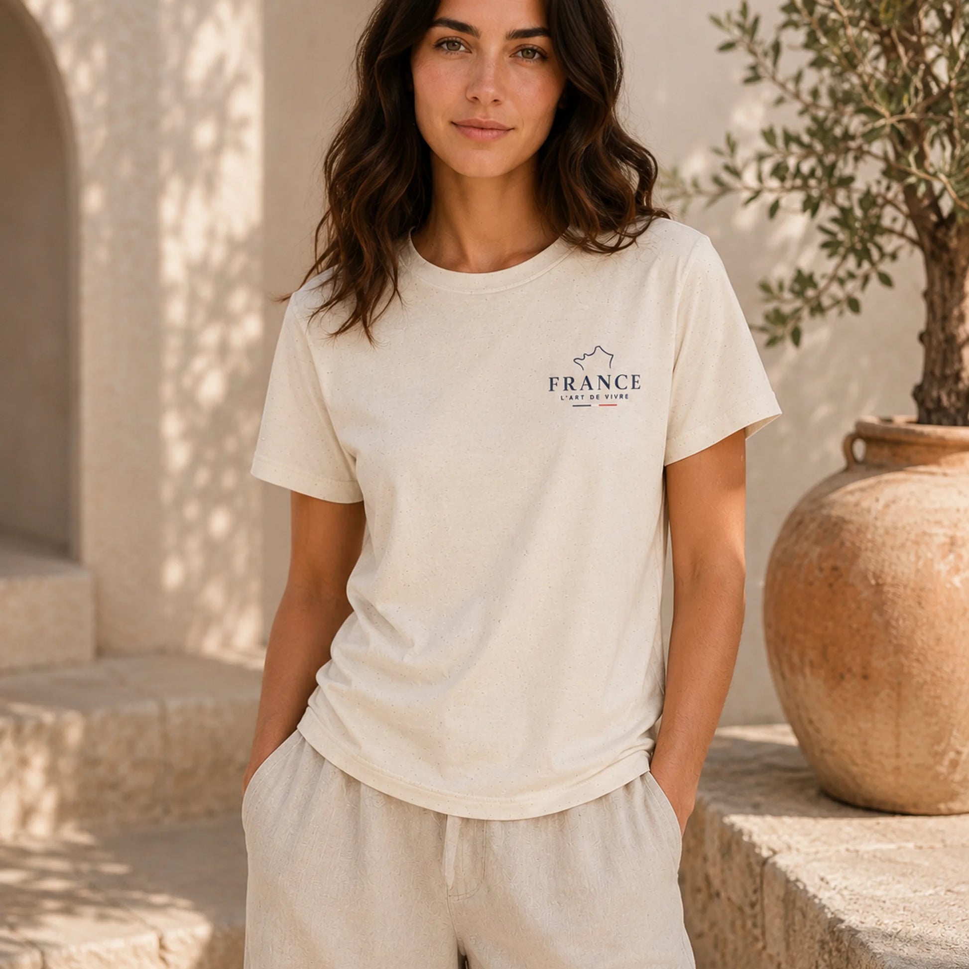 A woman wearing a cream-colored short-sleeve t-shirt with a small dark blue 'FRANCE' graphic on the left chest, paired with matching cream pants, set against a warm, sunlit stone patio background.