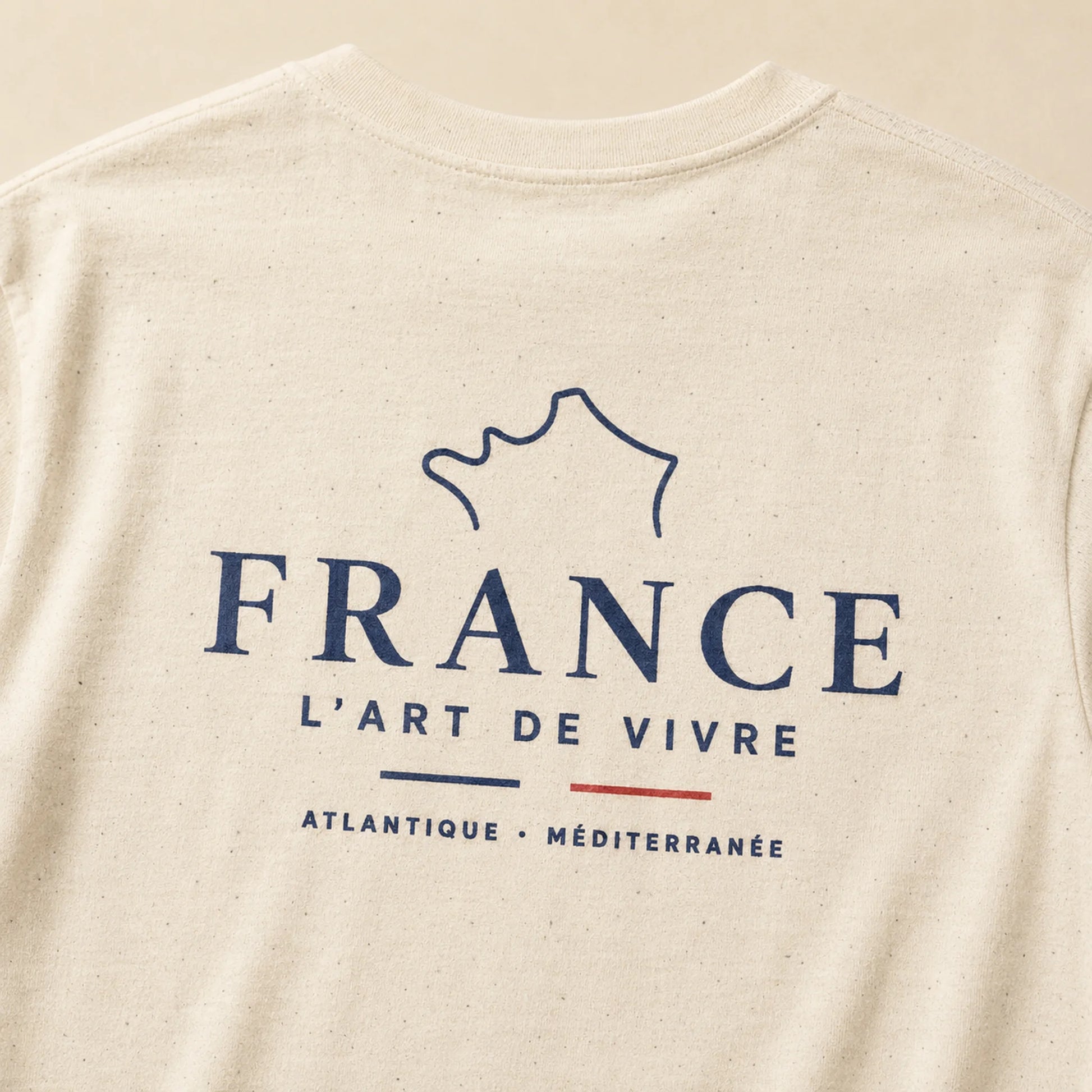 Close-up of a light beige speckled t-shirt featuring a navy blue graphic of the outline of France with the text 'FRANCE L'ART DE VIVRE' and 'ATLANTIQUE · MÉDITERRANÉE'.