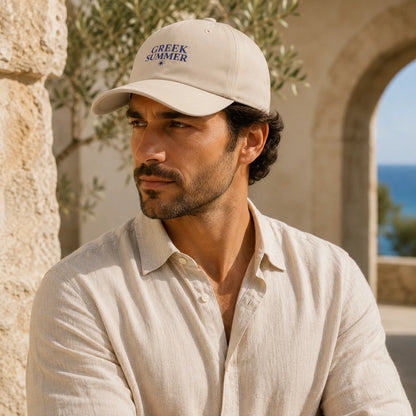 TP HERO: A man wearing a beige baseball cap with 'GREEK SUMMER' embroidered in navy blue, paired with a matching light beige linen shirt, set against a warm stone background with an archway and blue sea in the distance.