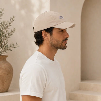 A man in profile wearing a light beige baseball cap with a small embroidered logo and a plain white t-shirt, standing in a warm, sunlit architectural setting with beige walls and a potted plant.