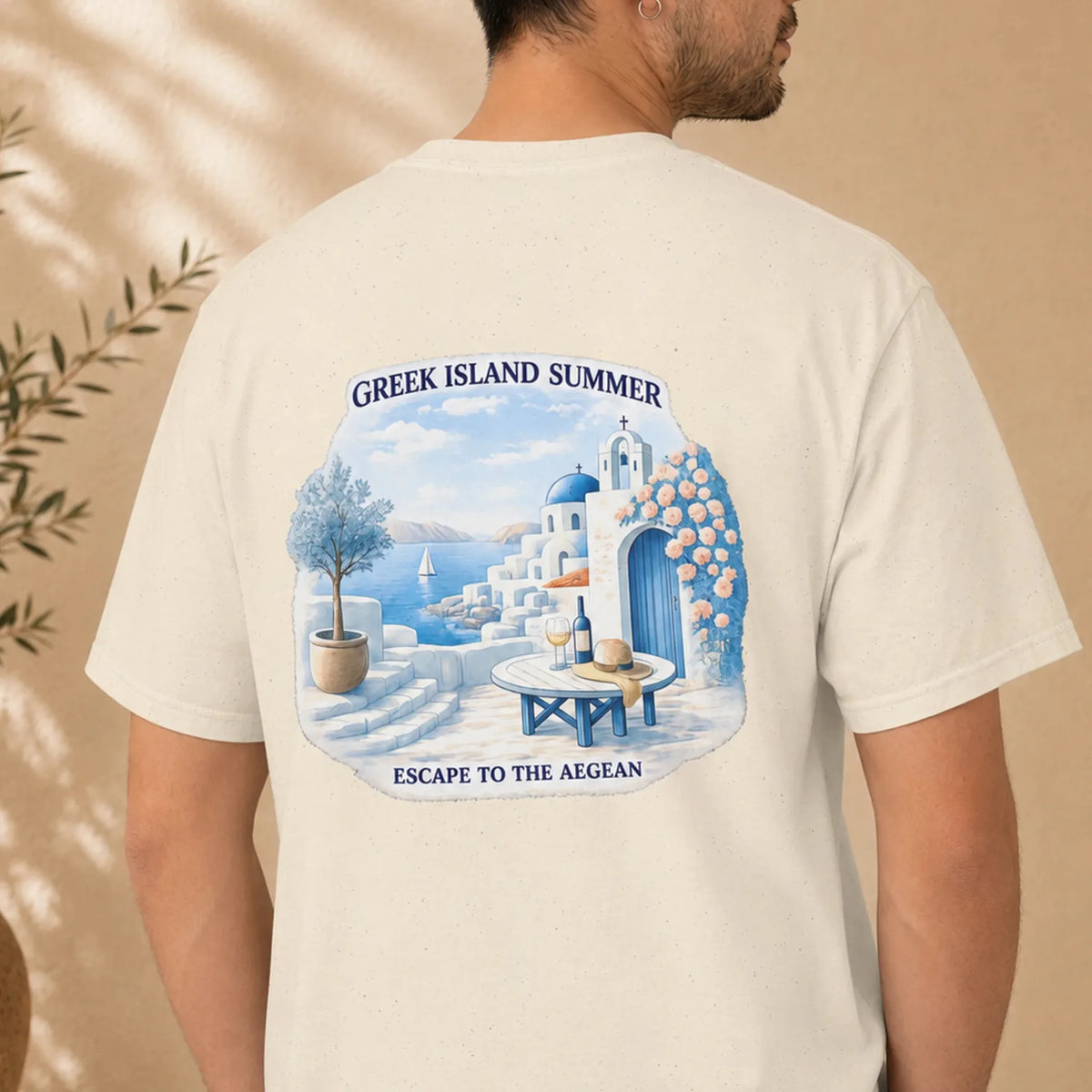 TP HERO: A man wearing a cream-colored t-shirt, viewed from the back. The shirt features a large graphic print of a Greek island summer scene with white buildings, blue domes, a blue table with wine bottles, and the text 'GREEK ISLAND SUMMER' and 'ESCAPE TO THE AEGEAN'.