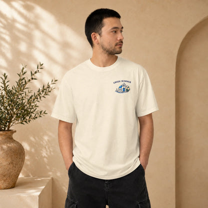 A man wearing a cream-colored crewneck t-shirt with a small blue and white graphic on the left chest, standing in a warm, beige-toned interior setting with a potted plant in the background.