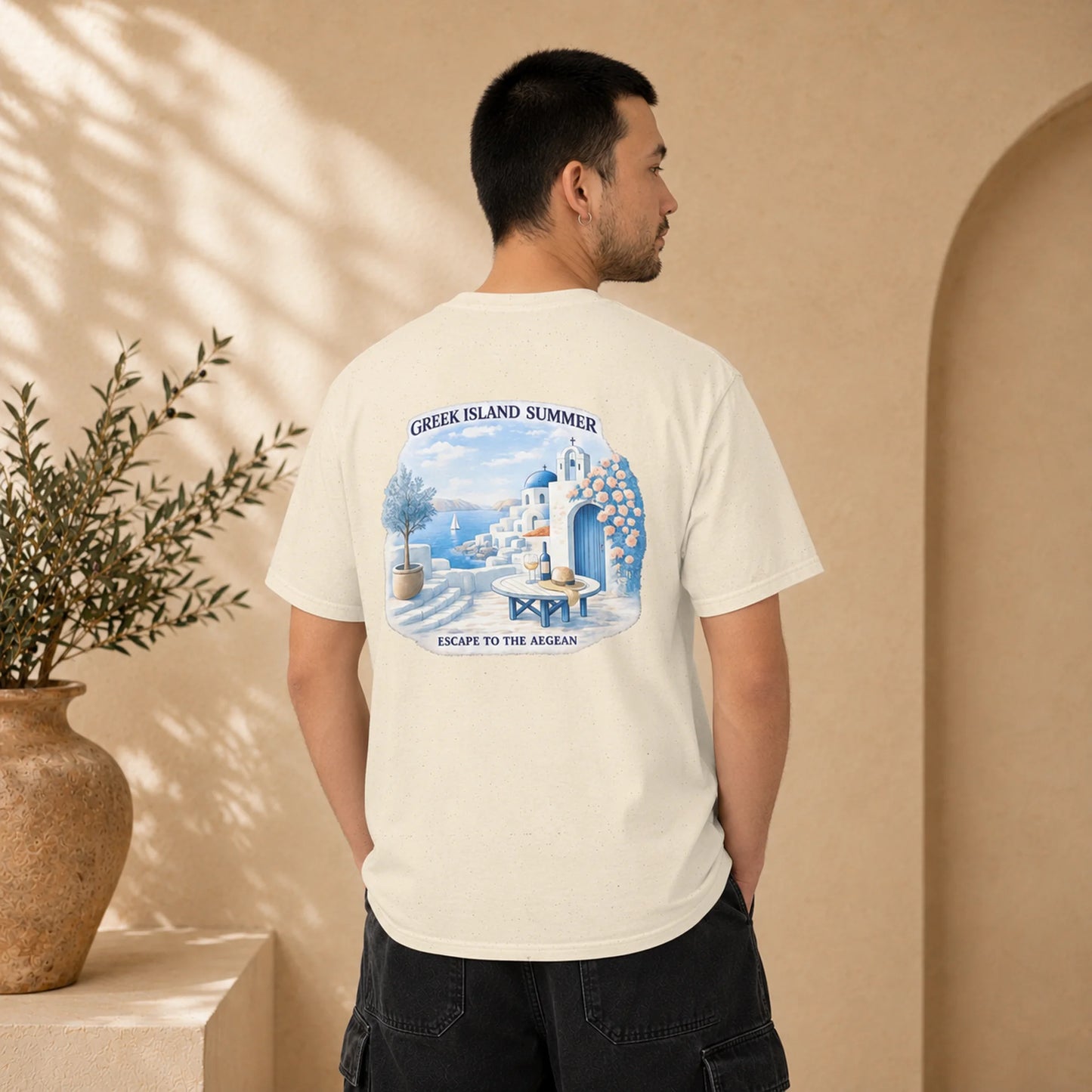 A man wearing a cream-colored t-shirt, viewed from the back. The shirt features a large graphic print of a Santorini-style Greek island summer scene with white buildings, blue domes, and a blue arched doorway, accompanied by the text 'GREEK ISLAND SUMMER' and 'ESCAPE TO THE AEGEAN'.