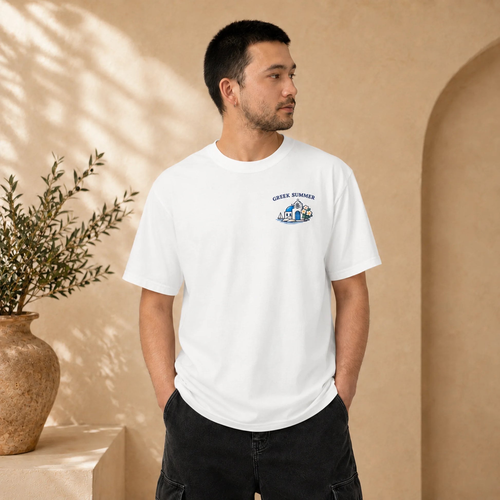 A man wearing a white crewneck t-shirt with a small blue and black graphic on the left chest that reads 'GREEK HOMER', standing in a warm beige architectural setting with a potted plant.