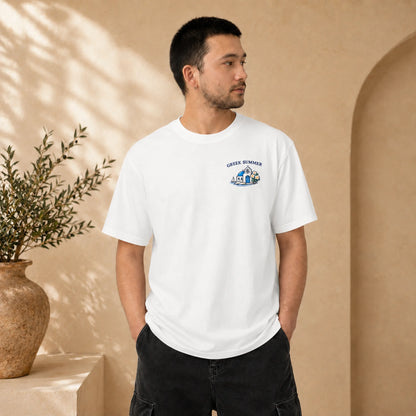 A man wearing a white crewneck t-shirt with a small blue and black graphic on the left chest that reads 'GREEK HOMER', standing in a warm beige architectural setting with a potted plant.