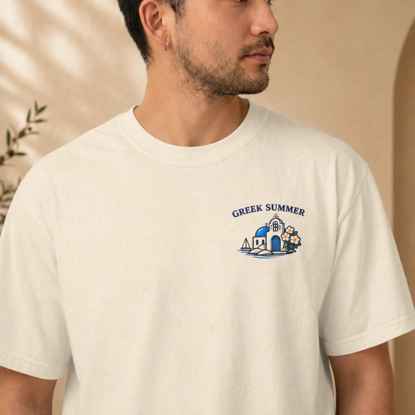 A man wearing a cream-colored t-shirt with a blue and white graphic on the left chest that reads 'GREEK SUMMER' above a small illustration of a building, sailboat, and flowers.