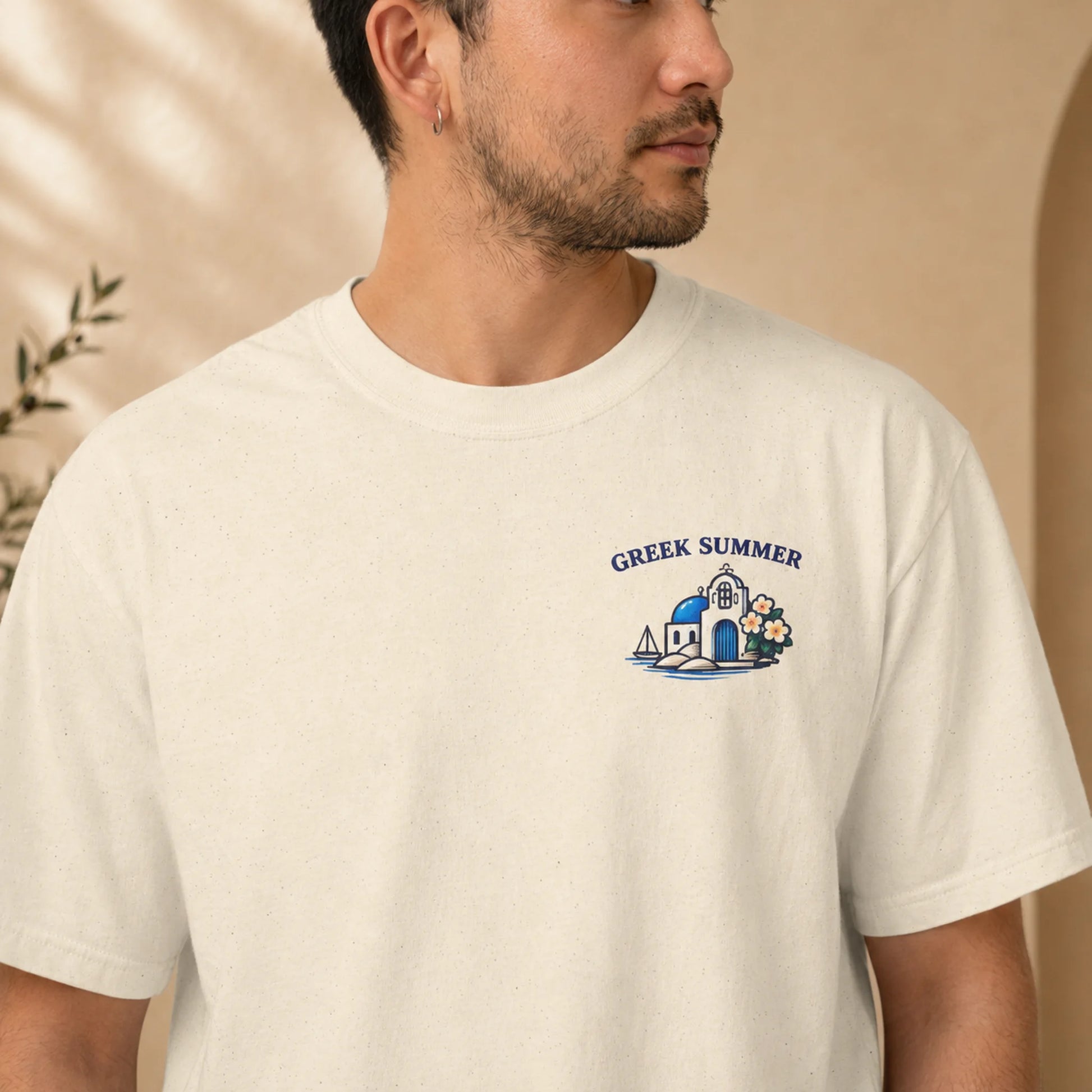 A man wearing a cream-colored t-shirt with a blue and white graphic on the left chest that reads 'GREEK SUMMER' above a small illustration of a building, sailboat, and flowers.