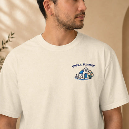 A man wearing a cream-colored t-shirt with a blue and white graphic on the left chest that reads 'GREEK SUMMER' above a small illustration of a building, sailboat, and flowers.