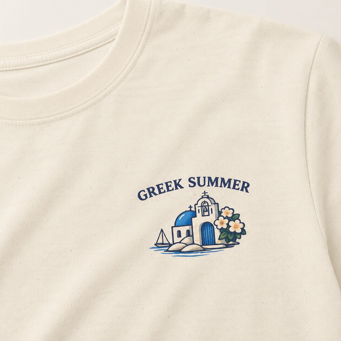 Close-up of an off-white crewneck t-shirt featuring a blue and white graphic of a Greek island church, blue domes, a sailboat, and white flowers, with the text 'GREEK SUMMER' printed above.