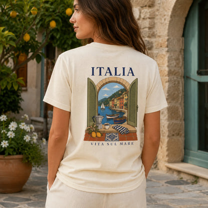 A woman wearing a cream-colored t-shirt with a large graphic print of an Italian coastal scene on the back, standing outdoors in front of a stone building.