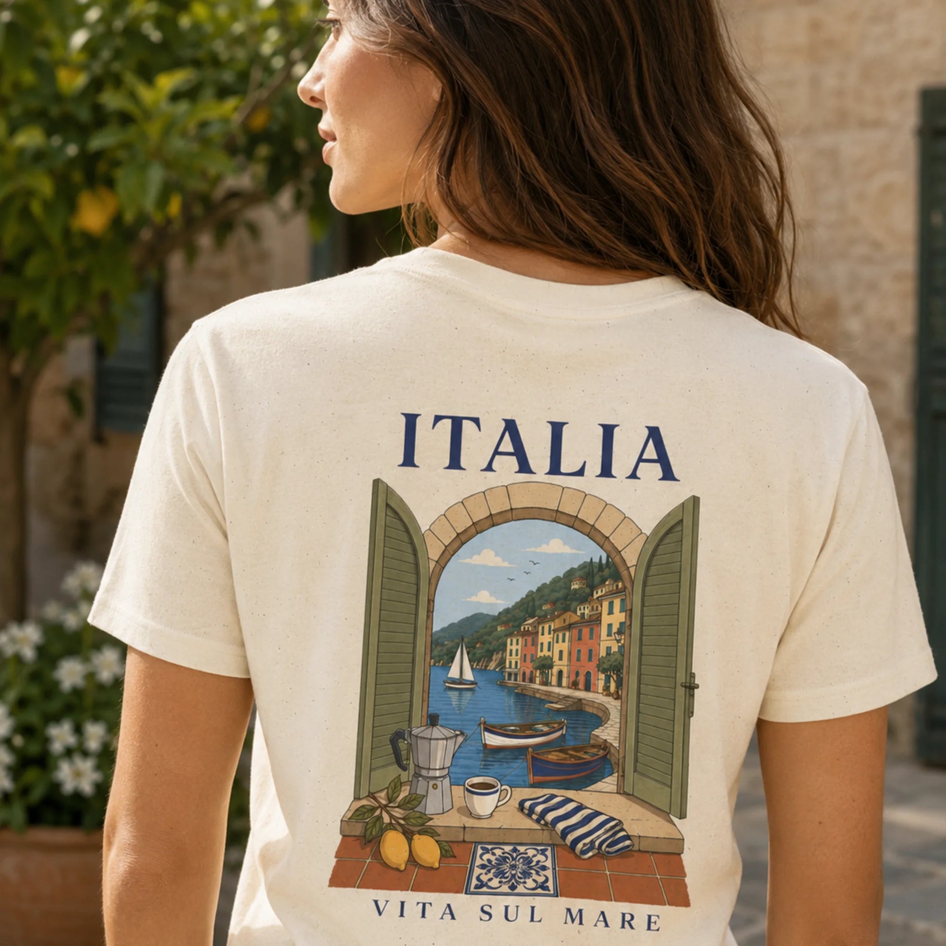 TP HERO: A woman shown from behind wearing a cream-colored t-shirt with a large graphic print of an Italian coastal town seen through an open shutter window. The text 'ITALIA' is printed above the graphic, and 'VITA SUL MARE' is printed below.