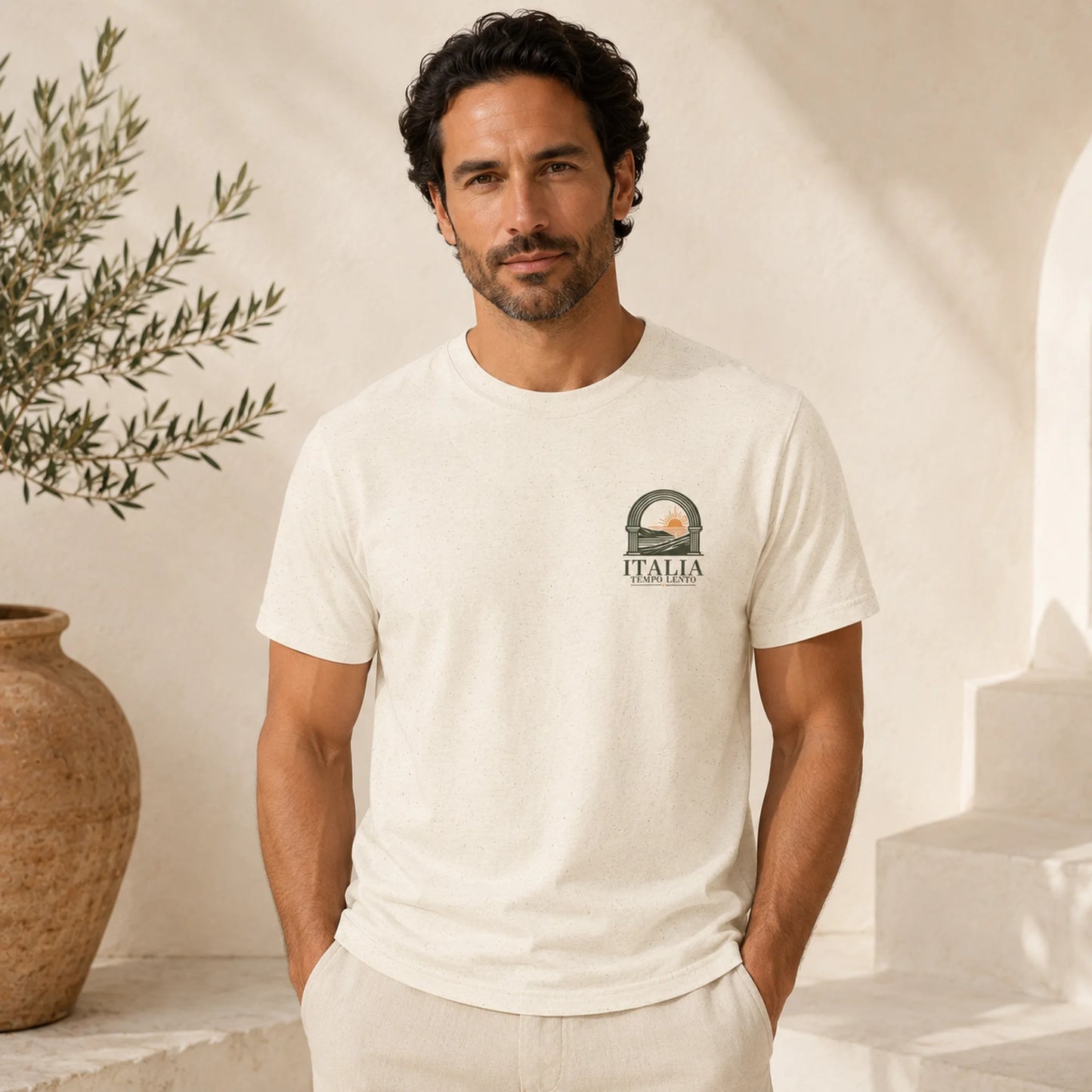 A man wearing a cream-colored short-sleeve t-shirt with a small graphic on the left chest that reads 'ITALIA' under a circular emblem, posed in a bright, sunlit room with a terracotta pot and white steps in the background.