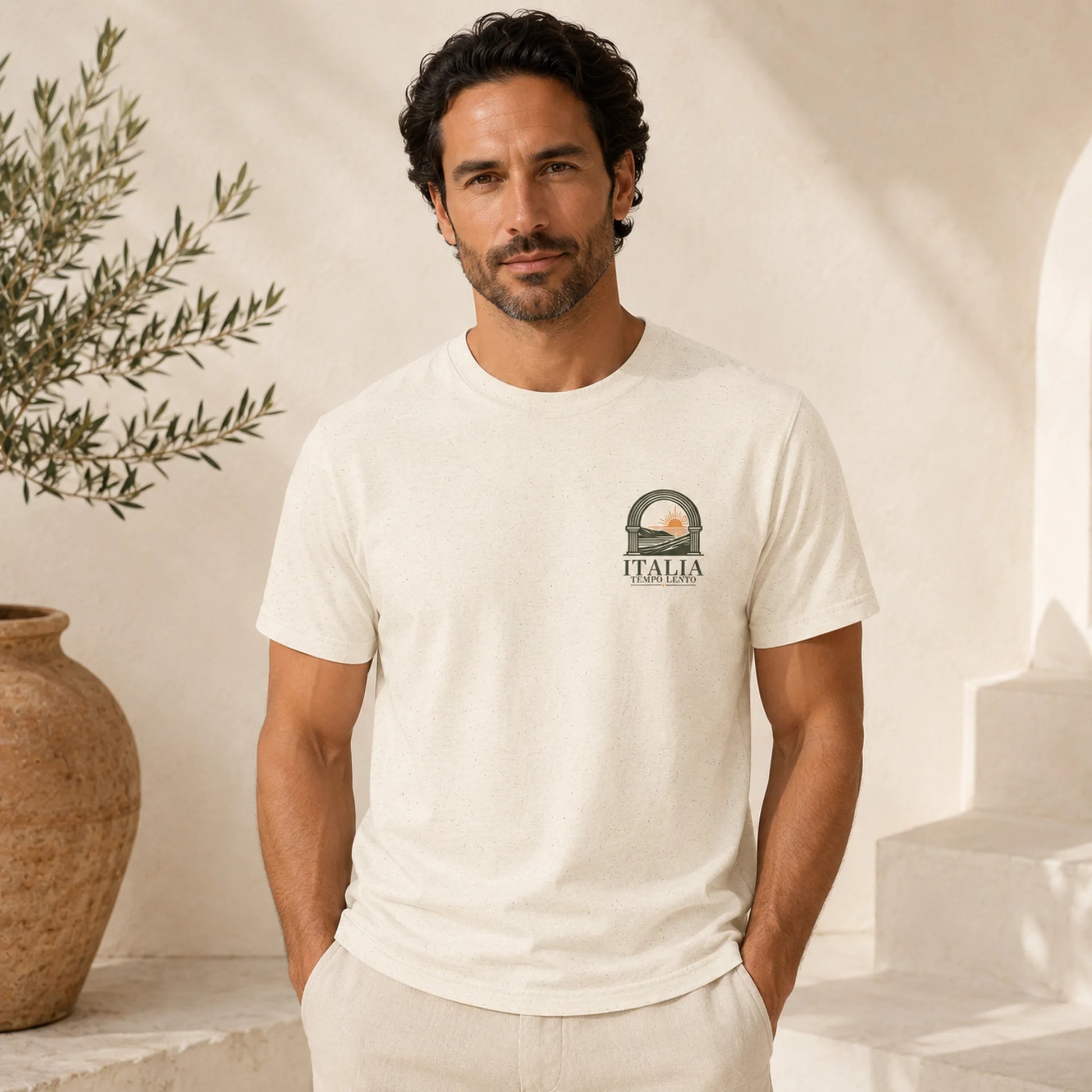 A man wearing a cream-colored short-sleeve t-shirt with a small graphic on the left chest that reads 'ITALIA' under a circular emblem, posed in a bright, sunlit room with a terracotta pot and white steps in the background.