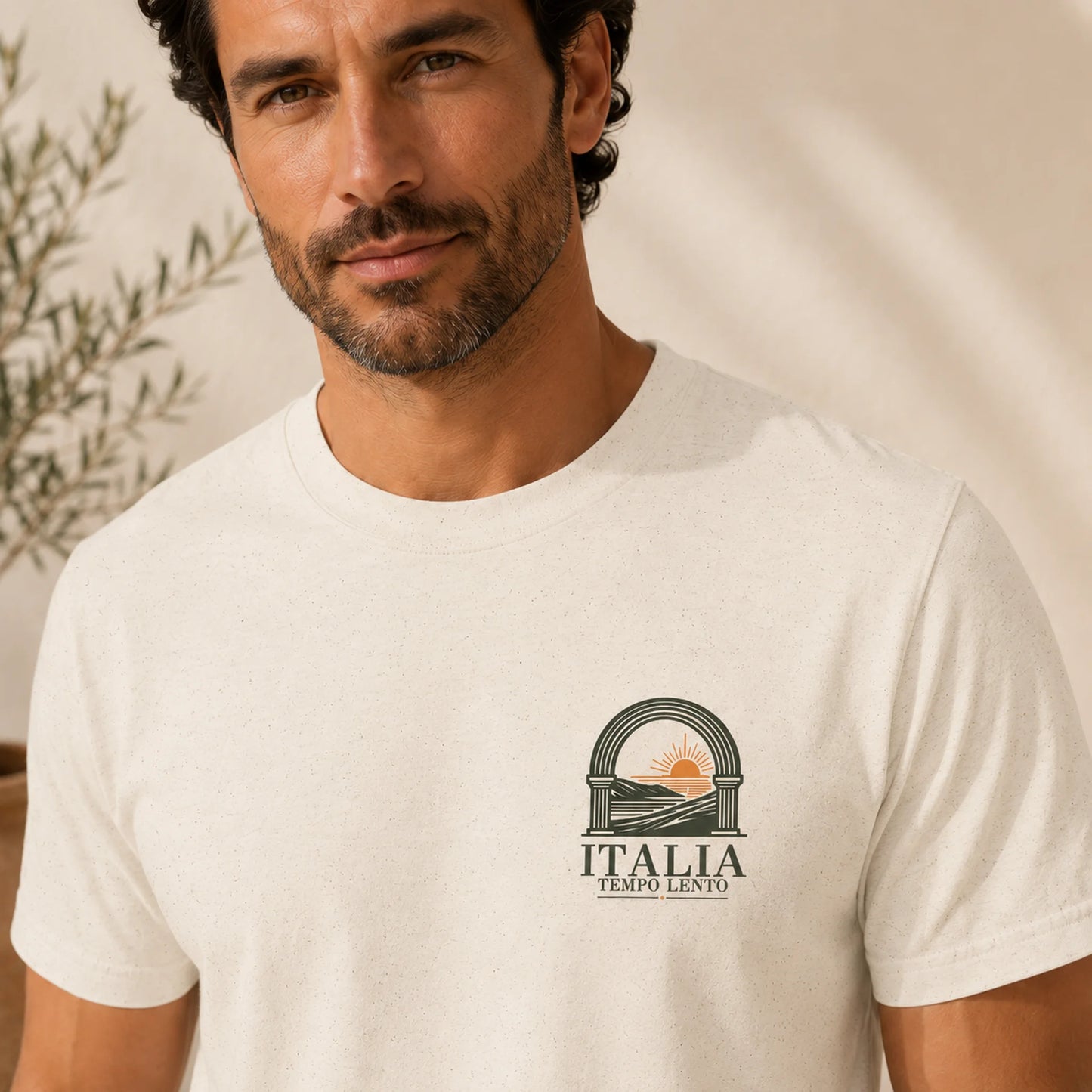 A man wearing a light cream-colored crewneck t-shirt with a small graphic on the left chest. The graphic features an archway with a rising sun and the text 'ITALIA TEMPO LENTO'.