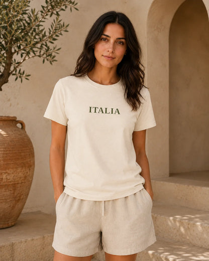 A woman wearing a cream-colored short-sleeve t-shirt with 'ITALIA' printed in dark green across the chest, paired with matching cream linen shorts, standing in front of a rustic beige stucco wall with an olive tree and a large terracotta pot.