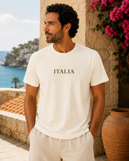 A man wearing a cream-colored crewneck t-shirt with the word 'ITALIA' printed in green across the chest, standing outdoors next to a stone wall with pink flowers and a view of the sea in the background.