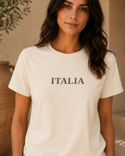 A woman wearing a cream-colored crewneck t-shirt with the word 'ITALIA' printed in dark green serif letters across the chest, set against a warm, neutral indoor background.