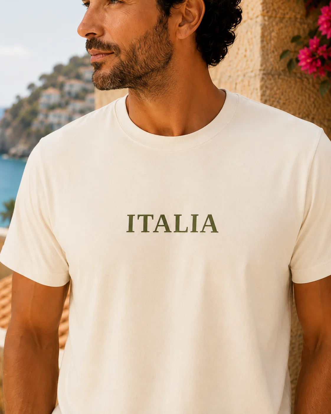 TP HERO: A man wearing a cream-colored crewneck t-shirt with the word 'ITALIA' printed in dark olive green serif letters across the chest, set against a warm Mediterranean background.