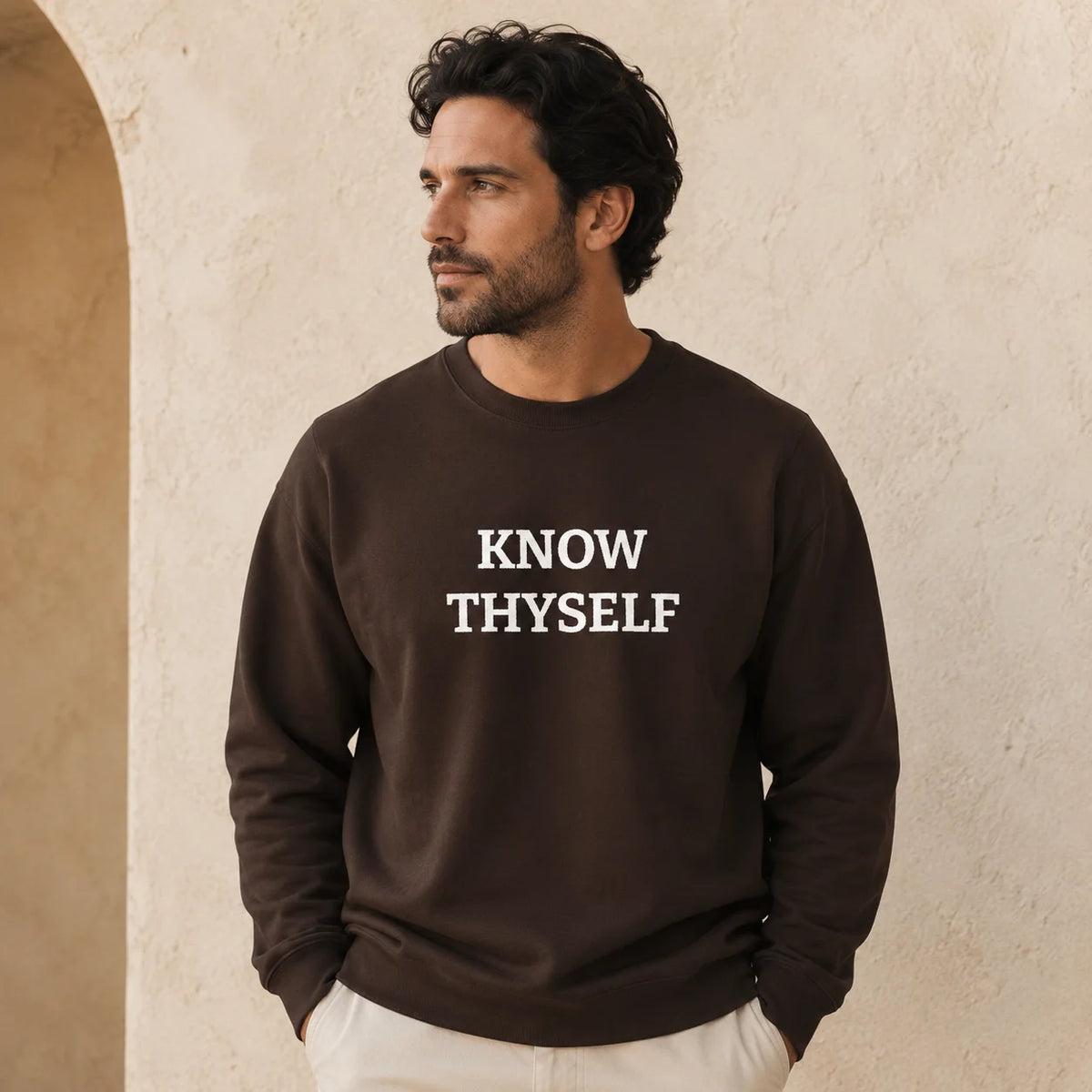 A man wearing a dark brown crewneck sweatshirt with the words 'KNOW THYSELF' embroidered in white block letters on the chest, standing against a textured beige stucco wall.
