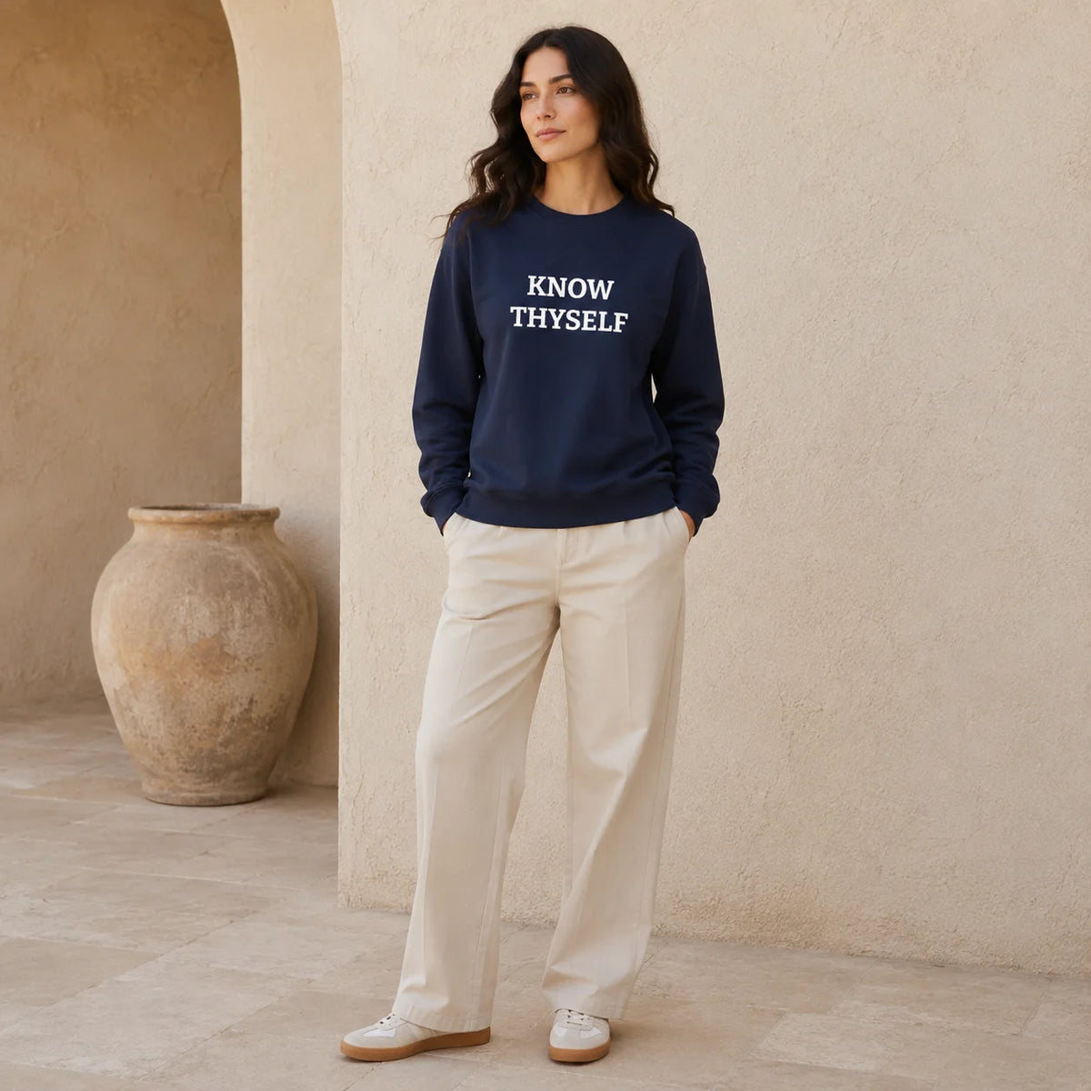 A woman stands in a warm, rustic architectural setting wearing a navy blue crewneck sweatshirt with the text 'KNOW THYSELF' embroidered in white across the chest, paired with cream-colored pants and white sneakers.