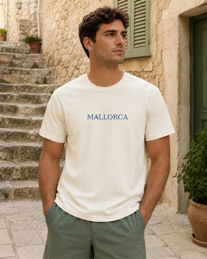 A man wearing a cream-colored t-shirt with 'MALLORCA' printed in blue across the chest, standing outdoors in front of a stone building with steps.