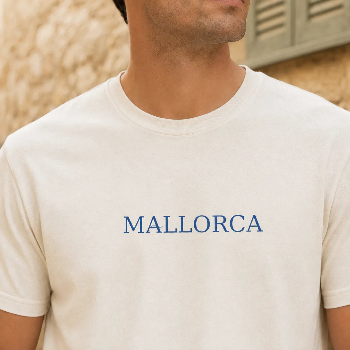TP HERO: A close-up of a man wearing a cream-colored crewneck t-shirt with the word 'MALLORCA' printed in blue across the chest, set against a warm, blurred background.