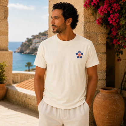 A man wearing a cream-colored short-sleeve t-shirt with a small blue and red geometric flower logo on the left chest, standing on a sunny stone terrace with a coastal ocean view in the background.