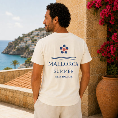 A man wearing a cream-colored t-shirt with a blue and red graphic on the back, standing on a sunny stone terrace with a Mediterranean sea and cliffside town in the background.