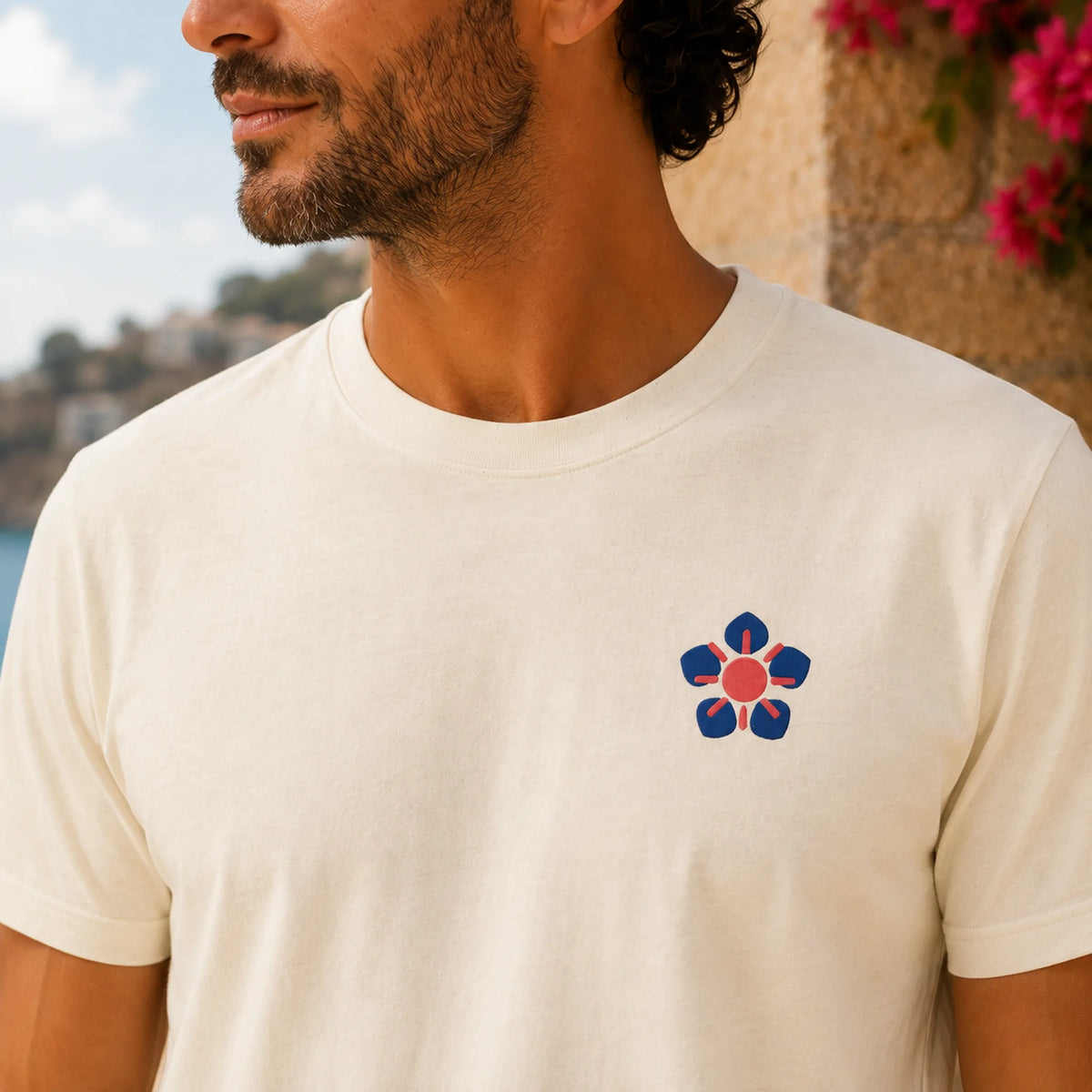 A man wearing a cream-colored crewneck t-shirt with a small stylized flower logo on the left chest in navy blue, red, and white, set against a blurred outdoor background.